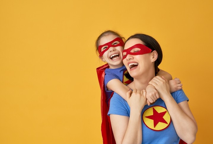woman & child in matching superhero costumes, child on her back, both smiling laughing, yellow background.